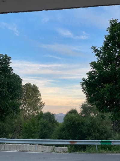 An exterior, eye-level, medium shot captures a scenic view of trees and mountains under a pale blue and orange sky. The foreground features a paved road and a concrete barrier with a metal guardrail, the latter showing two paint marks, one brown and one green. The middle ground is dominated by lush green trees of varying heights, with a few taller trees framing the edges of the shot. In the background, distant mountains are visible, softened by the haze and the fading light. The sky is a canvas of light blue streaked with thin, wispy white clouds, transitioning to a soft orange hue near the horizon. The overall impression is one of natural beauty, possibly a rural or mountainous area, with the road and guardrail suggesting a roadside viewpoint. The scene is devoid of people or any discernible human activity beyond the road infrastructure. The location is identified as Valkosel, Bulgaria.