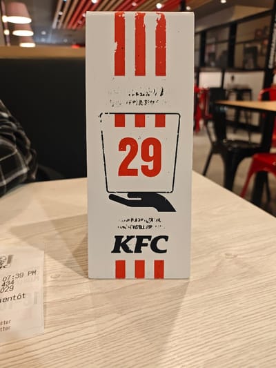 A vertical white cardboard stand, serving as a table number indicator, is positioned centrally on a light-colored wooden table surface within a KFC restaurant in Thionville, France. The stand features red vertical stripes at its top, a central graphic depicting a black-outlined bucket containing the large red number '29', a black hand icon positioned below the bucket, and the 'KFC' logo followed by three small red vertical stripes at its base. Faded, partially illegible text is visible above and below the bucket graphic.
To the immediate left of the stand, a white paper receipt is partially visible on the table, displaying the 'KFC' logo, a timestamp of '07:39 PM', the numbers '434' and '029', and the French word 'bientôt'.
A person wearing a black and white plaid shirt is partially visible on the left, seated at the table. Behind this individual, a dark-colored upholstered booth is present. The background, out of focus, reveals the interior dining area of the KFC restaurant, characterized by light-colored tables, a mix of black and red chairs, modern ceiling light fixtures with circular shades, and wooden ceiling slats.