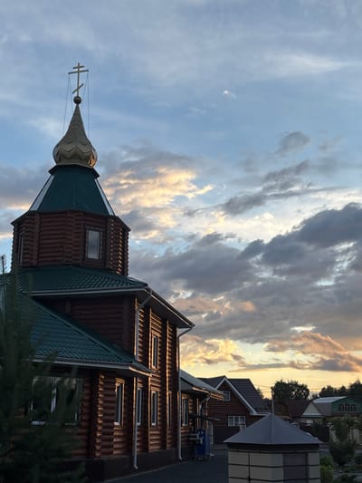 The image depicts the exterior of a wooden Orthodox church in the village of Surok, Russia, at dusk. The church features a green metal roof and a golden, octagonal dome topped with a cross. The building is constructed from horizontal logs, and several windows are visible along its side. The sky is filled with dramatic clouds, illuminated by the setting sun, creating a warm glow. In the background, other residential buildings and trees can be seen. A evergreen tree is partially in the foreground on the left side of the image.