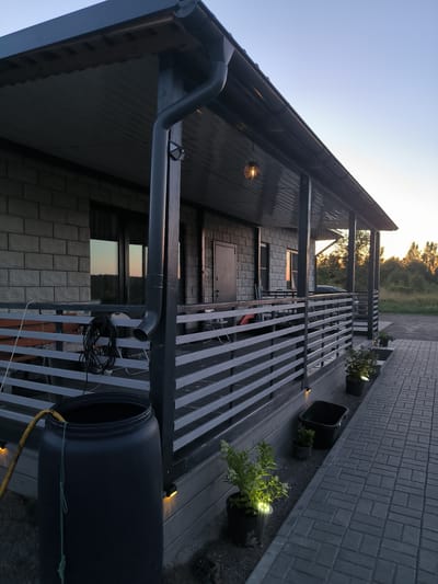 The image depicts the exterior of a modern, single-story house with a covered porch in Mezhozernoe, Russia. The house is constructed with light-colored stone or brick. A dark-colored porch extends along the front of the house, supported by dark posts. A railing with horizontal slats encloses the porch. A large black plastic barrel with a yellow hose is situated near the porch's edge, and a green rope hangs from it. Several potted plants are placed along the porch and the paved walkway, with some illuminated by small, upward-facing lights. The walkway is made of gray paving stones. A gutter system is visible along the roofline, with a downspout near the large barrel. The sky is clear and shows the soft light of either sunrise or sunset. The surrounding environment appears to be a rural or semi-rural area with trees and vegetation visible in the background. No people are present in the scene.
