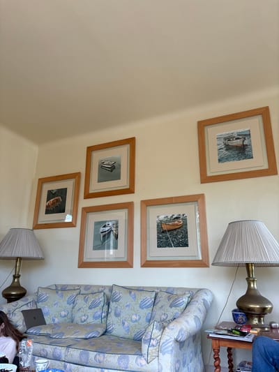 The image shows a living room interior with five framed pictures of boats on the wall. Two lamps, one with a white shade and the other with a gray shade, are on side tables beside a couch.  The couch is covered in a floral patterned fabric with three throw pillows. A laptop computer, a book, and a cup rest on the couch. The room is painted in a light beige color. The location of the room is in Dinard, France at the coordinates 48.638, -2.057.