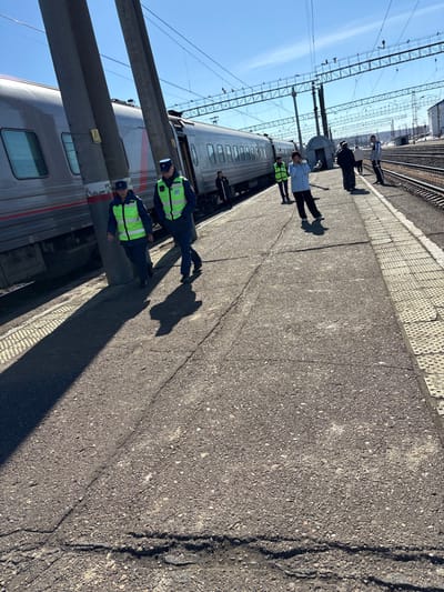 The image depicts a train station platform in Kropachyovo, Russia, under a clear blue sky. A silver passenger train, displaying a red Russian Railways logo, is stationary alongside the platform on the left. Two individuals in dark blue uniforms with bright neon-green reflective vests and dark caps are walking towards the foreground on the asphalt platform. Long shadows extend from them and from two prominent dark grey utility poles located between them and the train. One utility pole features a yellow triangular warning sign. Further down the platform, several other individuals are visible; one wears a light blue jacket and another a neon-green vest. The platform surface exhibits cracks and minor deterioration, with yellow tactile paving strips visible along its edge. In the background, multiple sets of train tracks are visible, crossed by an extensive overhead catenary system supported by metal gantries. Distant structures are faintly visible on the horizon to the right.