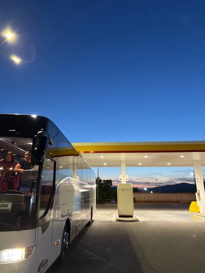The image shows a large bus parked at a Shell gas station at dusk in Kalotina, Bulgaria. The bus is white with a black windshield and front. The gas station has a yellow and white canopy. There are two illuminated light poles to the left of the bus. A mountain range is visible in the background. There are two people visible inside the bus.