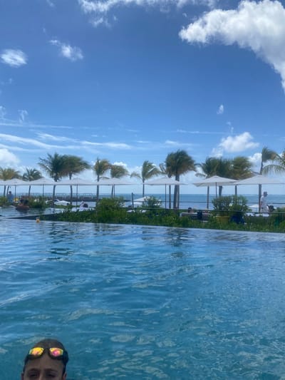 The image is a candid shot taken from a pool in Marina Maroma, Mexico. The pool water dominates the foreground, with a man's face visible in the lower right corner, wearing swimming goggles. Beyond the pool's edge, a row of palm trees and white umbrellas line a patio area. People can be seen relaxing and walking in this area. The horizon line is slightly visible in the background, with a glimpse of the ocean. The sky occupies the left side of the image, featuring a bright blue sky with scattered white clouds. The photo is oriented vertically.