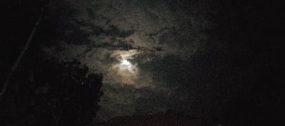 The image captures a nocturnal scene in South Kasipul ward, Kenya. The central subject is a bright, circular light source, consistent with the moon, partially obscured by dark, broken cloud cover located in the upper-middle to right section of the frame. The sky surrounding this area is black. In the lower-left quadrant, a dark silhouette of tree foliage is visible. A dark, linear object, likely a utility pole or similar structure, extends diagonally from the upper-left corner. The scene is devoid of human presence, discernible actions, or interactions. The image exhibits significant photographic noise, characteristic of low-light conditions.