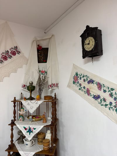 This is an indoor scene in Ust'-Kabyrza, Russia, likely a room within a traditional dwelling or museum. The environment is sparsely decorated with white walls and a wooden shelf unit. On the walls are two pieces of embroidered fabric: one with red floral patterns hangs to the left, and another with colorful floral patterns runs diagonally across the right. A dark wooden cuckoo clock hangs on the wall above the embroidered fabric on the right.

The wooden shelf unit displays various decorative objects. The top shelf holds an oil lamp with a green base and glass chimney, a candle, a small egg in a stand, a small wooden box, and a glass container with a rooster figurine. The second shelf features a doll in a colorful dress, a basket filled with colorful objects, a small bird figurine, and another figurine resembling an angel. The third shelf displays a metal object, a teapot with a floral design, a round wooden container with a patterned surface, and a standing figure made of what appears to be white yarn or fabric. A piece of embroidered fabric with a floral design is draped over the third shelf.
