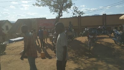 The image shows a scene in Kayna, Democratic Republic of the Congo, with a group of people standing on a dirt road in front of a tall beige wall. The road is mostly dirt, with some gravel and debris present. The wall is topped by a metal fence. There is a large tree in the foreground, which appears to be in front of a building with a red sign that is not entirely visible. The building has a large, open doorway.  The people in the scene are all adult males and appear to be casually interacting.  Four motorcycles are parked in the foreground, a fifth motorcycle partially visible behind them.  Several people are standing by and around the motorcycles. The motorcycles are all in good condition.  The scene is lit by the sun, and the sky is blue with some white clouds.