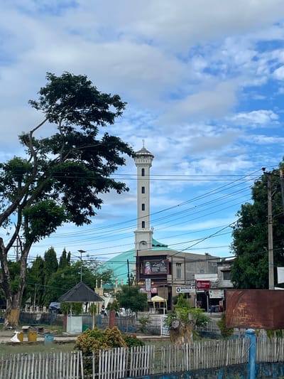 The image depicts a street scene in Pasar Paloh, Indonesia, with a prominent white minaret of a mosque rising against a blue sky with scattered clouds. A large tree with dark green foliage is situated in the left foreground, partially obscuring the view of the mosque. In front of the mosque, several low-rise buildings are visible, including shops with signage. A turquoise-colored dome of the mosque is partially visible behind these structures. A white picket fence runs along the bottom of the image, separating a grassy area with some bushes from the street. Electrical wires are strung across the scene, supported by wooden poles. The overall impression is one of a residential or commercial area with a significant religious landmark.