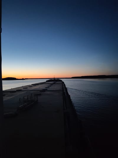The image shows a panoramic view of a large concrete structure extending into a body of water. The sky above is a gradient of deep blue at the top, transitioning to lighter blues and then a soft orange and yellow hue near the horizon, indicative of either dawn or dusk. A dark silhouette of land with trees forms the horizon line. The water is calm with subtle ripples and a faint wake trailing from the right side of the frame. The concrete structure, which appears to be a pier or breakwater, takes up the majority of the foreground and midground on the left side of the image. It is flat and wide, with a series of dark, rectangular objects arranged linearly along its surface. Some metal structures, possibly railings or equipment, are visible on the concrete. The overall scene suggests an outdoor environment, likely a port or waterfront area in Irdomatka, Russia, during the twilight hours. There are no people or visible animals in the frame. The lighting is dim, with parts of the concrete structure cast in deep shadow.