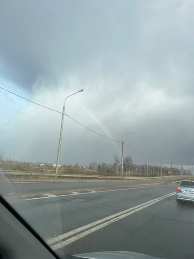 The image shows a roadside view from within a vehicle, possibly in Ivashevo, Russia. A faint rainbow arches across a gray sky. The road is wet with double white lines marking lanes. Streetlights line the road's edge with bare trees in the background. A silver car is visible in the adjacent lane. The scene is overcast.