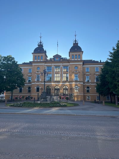 The image shows a large, ornate, tan-colored building with two distinct towers topped with dark domes and intricate spires, against a clear blue sky. The building features numerous windows, some arched and some rectangular, and decorative architectural elements. In front of the building is a circular garden area with flowering plants and a lamppost. A paved walkway and road lead up to the entrance of the building, which has a set of stairs. Two large trees flank the building. The scene appears to be a public square or plaza in the city of Oulu, Finland. The building is likely the Oulu City Hall. There are no people visible in the image. The lighting suggests it is daytime, likely morning or late afternoon given the long shadows cast.