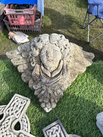 The image depicts a weathered, stone angel face sculpture lying on artificial green turf outdoors. Adjacent to the sculpture are stone Celtic crosses, also resting on the turf. In the background, there are crates and a folding chair. The location is Chirk, United Kingdom.