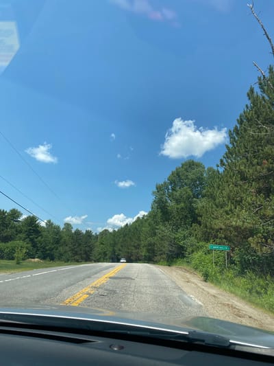 The image is a first-person view from a vehicle driving on a two-lane paved road under a bright blue sky with scattered white clouds. The road has a broken yellow center line and white lane markings. Dense green trees line both sides of the road, with taller coniferous trees on the right. A green street sign on the right side of the road reads "ST-ANDRÉ-AVELLIN 5". A white car is visible in the distance on the road. The foreground shows the dashboard and windshield of the vehicle. The environment appears to be rural, possibly in the region of Saint-André-Avellin, Canada, given the street sign.