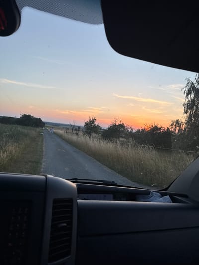 The image depicts a sunset over a rural landscape in Oberwetz, Germany. The viewpoint is from inside a vehicle, looking out through the windshield. The sky is a vibrant gradient of orange, pink, and blue, with wispy clouds. A paved road stretches into the distance, bordered by grassy fields and trees. In the middle distance, two figures appear to be walking along the road, accompanied by a dog. The overall mood is serene and peaceful, evoking the calm of a rural evening. The setting appears to be a country road, possibly in a field or farmland, bathed in the warm glow of the setting sun. The time of day is clearly sunset, indicated by the colors of the sky. The weather appears to be clear, with no visible rain or heavy clouds. The prominent objects include the road, trees, grass, sky, and the interior of the vehicle. There is no visible text in the image.