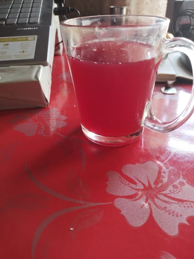 A close-up, eye-level shot shows a clear glass mug filled with a bright red liquid, possibly juice or tea, sitting on a red table with a subtle floral pattern. To the left of the mug is the corner of a silver laptop, partially obscuring a white and grey box beneath it. In the background, a metallic cylindrical object and other indistinct items are visible. The lighting suggests an indoor setting. The scene is located in Ikkiaul, Uzbekistan.