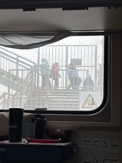 The image depicts a view from inside a train compartment in Barabinsk, Russia, looking out through a window onto a snowy outdoor scene. The foreground shows the interior train environment: a dark, crumpled fabric, likely a curtain or blind, is pulled across the top of the window frame. On a surface below the window, various objects are present: two dark travel mugs (one bearing the white text "ДА, НО НЕТ"), a light-colored cup, a metal bowl containing a spoon, a red plastic lid or container, and a silver fork. A partial white information sign with black text and Braille is visible on the train's interior wall to the lower right.

The exterior view, observed through the window, reveals a metal pedestrian overpass structure, likely at a railway station, under overcast or foggy conditions. Snow covers the ground and parts of the structure. Four individuals are present on the overpass: one person in a light blue/teal puffy coat stands on the upper stairs to the left, facing right; another individual in a red jacket stands behind them on the stairs. A third person in a dark jacket stands on a lower section of the stairs or landing to the right. A fourth individual, dressed in dark clothing, is seated or squatting on a low landing or bench near the right edge of the frame. A metal fence or barrier runs across the scene, featuring a rectangular white sign with black Russian text that translates to "Exit to the city is carried out through the station building." Below this sign, a yellow triangular warning sign with black symbols (likely a person and a train) is visible.