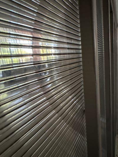 The image shows a close-up, angled view of a metallic roller shutter door, likely found on a shop or commercial building in Korsuntsi, Ukraine. The shutter is made of horizontal, ribbed metal slats that reflect light, creating a shiny, textured surface. To the right of the shutter, a vertical support structure, possibly a metal frame or pillar, is visible, also with a metallic or painted finish. The background reflections on the shutter suggest an outdoor environment with greenery and sunlight, indicating the door is likely situated on the exterior of a building. There are no people or discernible actions depicted in the image; it focuses solely on the architectural elements and their textures. The overall impression is one of a closed, secure entrance.