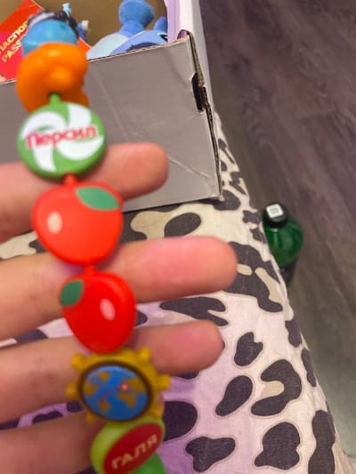 A close-up shot shows a hand holding a beaded bracelet. The bracelet features several charms, including a green disc with a white swirl and text that appears to be "Пероіл", two red apple-shaped charms with green leaves, a yellow gear-shaped charm with a blue globe in the center, and a green oval charm with red text that reads "ГАЛЯ". In the background, a cardboard box and a green bottle are visible. The overall scene is informal and likely captured indoors. No specific location in Vaganovo Massiv, Russia, can be determined from the provided image.