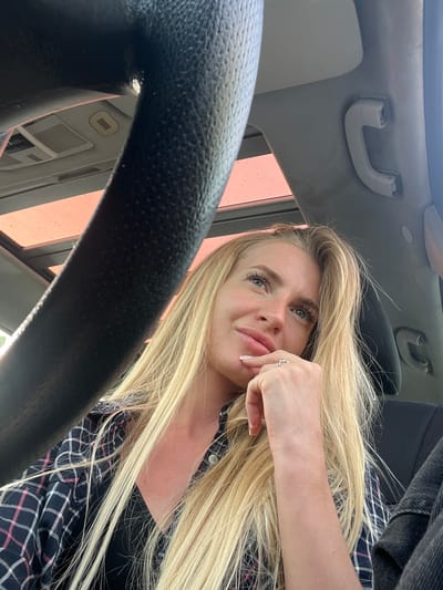 The image is a selfie taken from a low angle inside a car. The steering wheel is in the foreground. A blonde woman is visible, wearing a dark shirt with a patterned design. Her right hand is near her face, touching her lips. The interior of the car is visible, including the roof, a grab handle, and the side window. The location is Očinići, Montenegro.