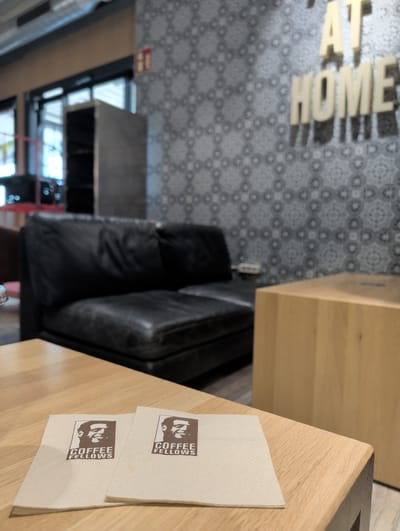This is a medium shot of a coffee shop interior. In the foreground, two paper napkins with the "COFFEE FELLOWS" logo are placed on a light-wood table. In the background, a black leather sofa is visible, with a patterned wallpaper and illuminated letters spelling "AT HOME" on the wall behind it. To the right, a light-wood cube-shaped structure is present. The overall ambiance suggests a comfortable seating area. The location is the Coffee Fellows branch in Lautzenhausen, Germany, situated within the airport.