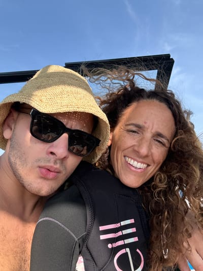 This image presents a close-up outdoor portrait of two individuals, an adult male and an adult female, in the city of Migdal, Israel. The scene is set under a clear blue sky, indicating daylight conditions.
The male, positioned on the left, wears a tan woven straw bucket hat and dark-framed rectangular sunglasses, obscuring his eyes. He has short stubble on his jawline and a portion of his bare chest and shoulder are visible, showing body hair. His lips are slightly pursed as he looks directly towards the camera. Sun patterns from his hat are visible on his face.
The female, positioned on the right, has tanned skin, voluminous, curly brown hair that appears wind-swept, and a broad smile revealing white teeth. A small reddish mark is visible on her forehead. She wears a black wetsuit or life vest adorned with a pink and light blue "BILLI" logo on the chest.
Both individuals are in close proximity, suggesting a shared moment. In the upper background, behind and above the subjects, a dark, angular metallic structure, possibly a frame from a boat canopy or watersports equipment, is visible against the clear sky.
