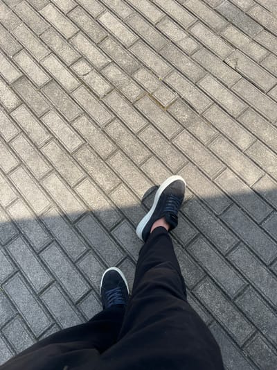 An overhead shot shows a person's legs and feet walking on a paved surface made of interlocking grey rectangular bricks laid in a herringbone pattern. The person is wearing dark pants and dark sneakers with white soles and dark blue laces. A shadow falls diagonally across the bricks from the upper left to the lower right of the frame, indicating a light source from the upper left. The scene appears to be an outdoor pedestrian area, possibly a walkway or sidewalk in Machara, Abkhazia.