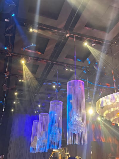 This detailed image captures the interior of an event venue or theater located in Ness Ziona, Israel. The scene is dominated by a complex overhead lighting and decoration arrangement against a dark ceiling.

The ceiling itself is dark, likely black or dark gray, with visible structural elements including metal beams and tracks from which numerous theatrical spotlights and stage lights are suspended. Integrated into the ceiling are several rectangular, lighter-toned panels. Numerous lighting fixtures are active, projecting distinct beams of light through an apparent atmospheric haze. Prominent light beams include a strong yellow-amber beam emanating from a fixture in the upper-middle left, another yellow-amber beam to its right, and several blue light sources across the ceiling, some projecting focused spots and others creating broader washes. One fixture near the upper-middle right exhibits a multi-colored, patterned light output.

Below the lighting grid, five decorative chandeliers are suspended. Four of these, arranged from largest on the right to smallest on the left, consist of clear or translucent cylindrical outer structures filled with numerous vertically hanging strands. Each cylinder encases a smaller, traditional crystal-style chandelier. These four chandeliers are primarily illuminated with vibrant blue and purple light, casting cool tones onto their strands and internal components. To the far right, a large, multi-tiered chandelier with ruffled or layered opaque white/off-white elements is partially visible, emitting a warm, soft glow.

In the background, dark stage curtains are visible along the rear wall, particularly on the left, where they are illuminated by blue stage lighting. The presence of visible light beams indicates a hazy or smoky atmosphere within the venue. At the very bottom center of the frame, the top of a head with dark, possibly curly, hair is partially visible, suggesting an individual is present and observing the scene from a low vantage point.