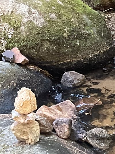 The image shows a close-up view of a cairn of stones stacked near a shallow stream. The cairn is composed of three rough, tan-colored stones. To the left of the cairn are larger rocks, one of which is covered in moss. The stream bed is visible with several stones submerged in the water. The background consists of more rocks and foliage. The scene is set in a natural, outdoor environment. The location of Gumières, France is not depicted or suggested by the visual information in the image.