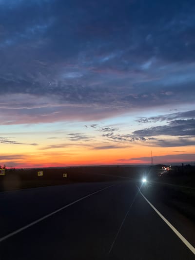The image depicts a highway at dusk, with the road stretching towards the horizon. The sky is a canvas of dramatic colors, transitioning from deep blues and purples at the top to vibrant oranges and reds near the horizon, indicating the setting sun. Wispy clouds are scattered across the sky, adding texture to the twilight scene. On the side of the road, there are a few yellow signs with black symbols. In the distance, a tall communication tower is visible, and further along the highway, headlights from oncoming vehicles create bright streaks of light. The overall scene suggests a moment of travel or transition as day turns into night. There is no discernible information about the city of Orlovo, Russia.