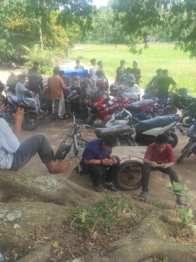 The image captures an outdoor gathering of several young men or boys in a semi-rural setting in Buketdalam, Indonesia. Numerous motorcycles and scooters are parked closely together in the midground and foreground, indicating a group arrival.
In the foreground, two individuals are seated on the ground near the large, gnarled roots of a prominent tree. One wears a red shirt and black pants, leaning against a motorcycle wheel while looking at a mobile phone. Another, in a dark blue shirt and black pants, is also seated, engaged with a phone; both have bare feet visible. Many other individuals are standing or sitting around the parked vehicles, some wearing traditional Islamic peci (kufi caps).
The background features lush green fields with a small, simple building visible in the distance. Dense trees and bushes line the edges of the scene, contributing to the natural, outdoor environment. The activity appears to be a casual social gathering or meeting. The time of day is bright daylight, suggesting clear weather. The overall atmosphere seems relaxed. While some text is visible on vehicle license plates, it is too indistinct to be clearly read.