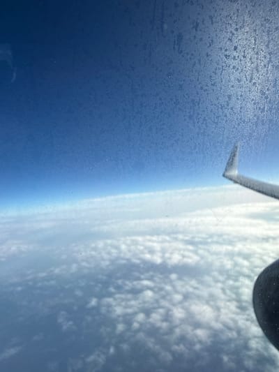The image depicts a view from an airplane window, showing a vast expanse of clouds below a clear blue sky. The airplane's wing is visible in the foreground, extending diagonally across the right side of the frame. The window itself has a slightly frosted or textured appearance, with some smudges or streaks visible. The scene is a typical aerial view, with no specific landmarks of Tihovići, Bosnia and Herzegovina identifiable. The focus is on the sky and the cloud layer.