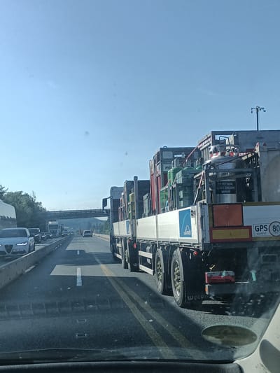 A sunny daytime view of a traffic jam on a highway in the city of Hrusice, Czechia. The camera is positioned inside a vehicle, looking forward through the windshield. In the foreground, the dark asphalt road is visible, with white and yellow lane markings. A white car with a license plate "2UA 3054" is in the left lane. Further ahead, several trucks and cars are stopped in traffic. The main focus is a large truck on the right, carrying several large green and silver gas cylinders. One cylinder has a label reading "UN 1977 DUSIK, HLUBOCE ZCHLAZENY KAPALNY". The truck also has a "Linde" logo on its side and a "GPS control 80 km" sign. The sky is clear and blue. In the background, a highway overpass crosses the road, and trees line the sides of the highway.