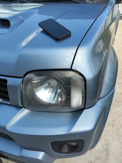 The image shows the front right corner of a light blue car, possibly a Suzuki Jimny, parked outdoors on a concrete surface. A black smartphone is resting on the hood of the car. The car's headlight is visible, with its internal reflectors catching the light. The hood features a prominent air intake scoop. The scene is brightly lit, suggesting it is daytime with clear weather. The background is slightly out of focus but appears to be an outdoor area with a light-colored fence or wall in the distance. There is no text visible in the image. The overall impression is a simple, everyday scene of a car parked outside.
