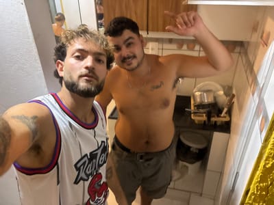 Two shirtless men pose for a selfie in a kitchen. One wears cargo shorts, a silver chain necklace, and has chest tattoos. The other dons a white Toronto Raptors basketball jersey. Behind them, the kitchen has tiled floors and walls, a wooden cabinet, and countertop items like cookware and eggs. The image is oriented upside down.