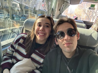 The image captures a selfie-style portrait of two young adults seated inside a vehicle, likely a bus or coach, during daytime in Aix-en-Provence, France. On the left, a female with long brown hair, wearing a striped top (maroon, light blue, and beige), smiles broadly at the camera. A white object rests on her lap. To her right, a male wears dark reflective sunglasses and a dark, zippered sweater with a strap across his chest; he looks directly forward with a neutral expression. Both are seated in light grey upholstered seats. The window to their left reflects the urban environment outside, showing bare trees, multi-story buildings, several parked cars (including a light blue vintage model), and street signs, notably a red circular 'No Entry' or 'Do Not Enter' sign. The scene is brightly lit by sunlight.