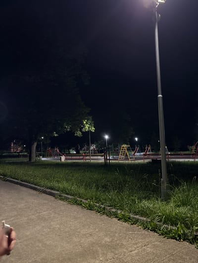 The image depicts a nighttime view of a children's playground in Pirdop, Bulgaria. A tall street lamp with a bright light illuminates the scene from the right side of the frame. The playground equipment, including slides and climbing structures, are visible in the background, with some lights from nearby buildings or streetlights casting a faint glow. The ground of the playground appears to be a mix of sand and grass. A concrete pathway runs along the bottom of the image, with a strip of grass and some weeds growing alongside it. The overall atmosphere is quiet and deserted, suggesting the playground is not in use at this time. A hand holding what appears to be a phone or small object is partially visible in the lower left corner of the frame.