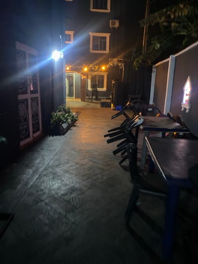 The image depicts an outdoor, paved area at night in Igbessa, Nigeria. The scene is illuminated by multiple artificial light sources, casting varying shadows and highlights.
On the left side, a dark-colored building features a bright, wall-mounted light fixture, emitting a strong glare. Below this, a rectangular planter box contains green foliage with small red or pink flowers. Adjacent to this, a white, paneled door or gate structure with black, ornate designs is visible.
The central portion of the image displays a concrete pathway with a repeating geometric tile pattern. This path leads towards an illuminated archway entrance in the background, suggesting an entry point into a building. Above this archway, string lights are visible. A dark-colored chair is positioned near the entrance. A single human figure, indistinct due to distance and low light conditions, is visible standing or walking in the background near the building's entrance. The building itself appears multi-storied, featuring several white-framed windows and a wall-mounted air conditioning unit on an upper level.
To the right, a low wall with a darker top runs parallel to the pathway. Embedded in this wall is a decorative niche or faux window, emitting a soft, colored light. In the right foreground, two dark-colored tables with blue legs and dark tops are present. Stacked horizontally on these tables are multiple dark-colored plastic chairs, indicating the area is either closed or being prepared for use. Above the right wall, dark green, broad-leafed foliage is visible against the night sky.