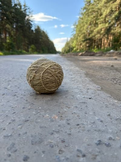 A ball of twine rests on a rough, grey asphalt surface. The twine is a natural, earthy color and is wound tightly into a spherical shape. The asphalt appears weathered and textured, with small pebbles and darker grey patches visible. To the right of the twine, a blurred path or road stretches into the distance, lined by trees with green foliage. The trees are out of focus, creating a bokeh effect. The sky above is a bright blue with scattered white clouds. The scene suggests an outdoor setting, possibly a rural road or path, given the natural materials and the presence of trees and sky. The lighting indicates it is daytime, with the sun casting light and creating shadows, though the shadows are not prominent. There is no visible text in the image. The overall impression is simple and natural, with a focus on the texture of the twine and the asphalt, and the blurred, natural background.
