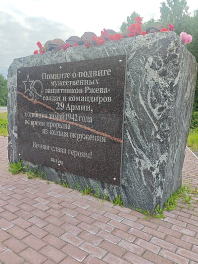 The image shows a monument in Esinka, Russia, dedicated to the memory of soldiers and commanders of the 29th Army who died in the winter of 1942 during a breakout from encirclement. The monument is made of granite and has an inscription in Russian. On top of the monument are several military helmets and artificial red and pink flowers. The monument is situated on a paved area with a brick walkway. The surrounding environment is natural, with trees and grass visible in the background. The sky is overcast.