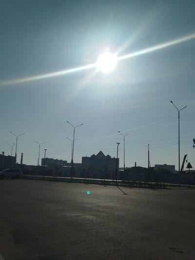 The image depicts an outdoor scene under a bright, slightly hazy sky with the sun intensely shining, creating a lens flare effect. In the foreground, a dark asphalt surface, likely a road or parking lot, is visible. A white SUV is parked on the left side of the image, facing away from the camera.
The middle ground shows a row of buildings, some appearing industrial or administrative, with a distinct minaret on the far left, suggesting a location in Kazakhstan, specifically Kokshetau, given the context provided. Several streetlights are positioned along the road. Construction cranes are visible in the background on the right, indicating ongoing development or industrial activity. The sky has a faint contrail. The overall impression is an urban or semi-urban environment on a sunny day.