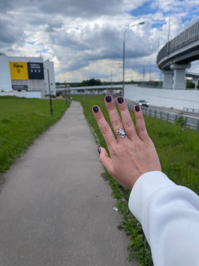 A hand with a silver ring on the ring finger, featuring a light blue stone and floral accents, is presented against an outdoor backdrop in what appears to be Razvilka, Russia. The hand is wearing dark-colored nail polish and is attached to a white garment, possibly a sweater or jacket. Behind the hand, a gray paved pathway stretches into the distance, flanked by green grassy areas. A modern building with large signage is visible on the left side of the frame. Above, a cloudy sky fills the upper portion of the image. A highway overpass is present on the top left of the image.