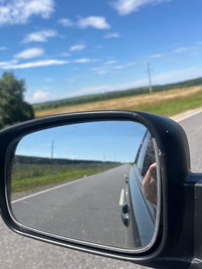 A close-up, slightly angled shot shows a car's side-view mirror, reflecting a bright blue sky with scattered white clouds. The mirror itself is rectangular and framed in black plastic. It is dirty with several smudges and small specks.
The reflection in the mirror reveals a rural landscape. A paved road stretches into the distance, flanked by green fields and some trees. A telephone pole is visible in the background, and a power line can be seen running across the frame. In the lower right portion of the mirror's reflection, a person's hand is visible.
The exterior of the car is partially visible outside the mirror. The environment appears to be outdoors on a sunny day. The image was taken from inside the vehicle, looking out. The scene does not provide specific information about the city of Kukshumy, Russia.