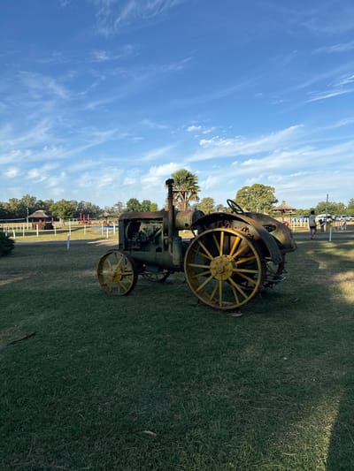 An antique tractor stands on a grassy field under a blue sky with wispy clouds. The tractor, primarily green with yellow wheels, shows signs of wear and rust. A steering wheel is visible. In the background, a fenced area with structures resembling small huts can be seen, along with some trees. A possible airplane is parked behind a distant fence. The scene appears to be an outdoor display at a rural or historical site.