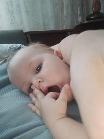 A close-up, low-angle, indoor shot shows a baby lying on a light blue, corduroy-like surface, likely a couch or bed. The baby's head is turned to the left, and their right hand is in their mouth. Their eyes are open and looking towards the viewer, with a slightly surprised or curious expression. The baby's mouth is open, and their lips are slightly red. The background is out of focus, but appears to be a wall with a textured pattern, possibly wallpaper, and a dark wooden shelf with what look like decorative stones on top. A sliver of a dark grey sofa armrest is visible in the upper left corner. The lighting suggests it is daytime, but the specific time of day is unclear. There is no visible text in the image.
