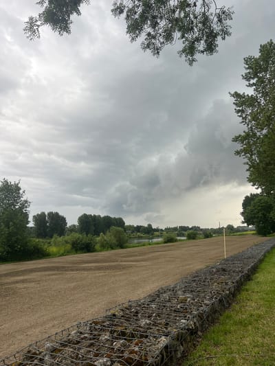 The image depicts a cloudy day with a dramatic sky filled with dark, ominous clouds. In the foreground, a gabion wall made of wire mesh filled with stones runs along a grassy area. Beyond the gabion wall is a large expanse of dry, tilled earth. Further into the distance, a line of lush green trees borders a body of water, likely a river, which is visible in the mid-ground. In the background, more trees and some buildings can be seen, with a bridge visible on the far right. The overall scene suggests a rural or park-like setting within the city of Venlo, Netherlands, possibly a riverfront area undergoing development or maintenance. There are no people or animals visible in the image.