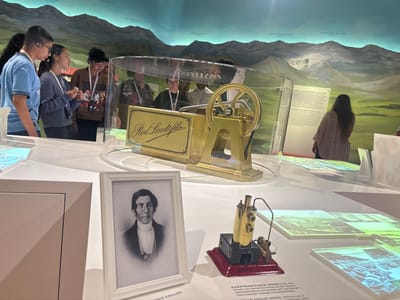 The image captures a scene inside a museum, likely the Lindt Home of Chocolate museum in Kilchberg (ZH), Switzerland, given the "Rod Lindt fils" display.

A group of young people are looking at exhibits. Some have audio devices. The displays include a gold-colored scale model under glass, a framed black-and-white portrait labeled "Louis Cailler," and a small steam engine model on a red base.

In the background, a mural depicts mountains with labels like "Schreckhorn" and "Gross Fiescherhorn," and text referencing Switzerland as the "land of chocolate." A woman is observing an information display.

The museum's setting is illuminated by bright, even lighting.