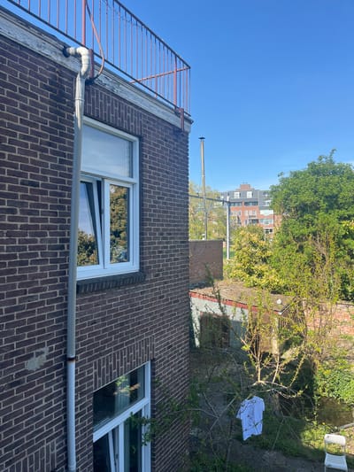 The image depicts the exterior of a brick building, possibly located in Maastricht, Netherlands, given the architectural style. The building features white-framed windows, some of which are open, reflecting the surrounding foliage. A grey drainage pipe runs horizontally along the brick wall. An orange metal railing is visible on the roof of the building.

In the background, a yard with trees, a building, and a white garment hanging on a tree can be seen. The sky is clear and blue.