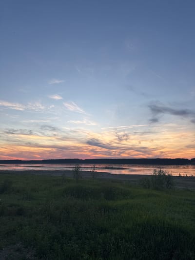 The image captures a serene sunset scene over a body of water, likely a river or lake, with a densely forested treeline in the background. The sky displays a gradient of colors, transitioning from a soft blue at the top to warm hues of orange and pink near the horizon, indicating the sun is setting. Wispy clouds are scattered across the sky, some catching the last rays of sunlight. In the foreground, a grassy area slopes down towards the water, with a few sparse bushes and tufts of grass visible. The water's surface reflects the colors of the sky, creating a shimmering effect. The overall atmosphere is tranquil and peaceful. This scene is located in Dolno Linevo, Bulgaria. There are no people or specific actions depicted in the image.