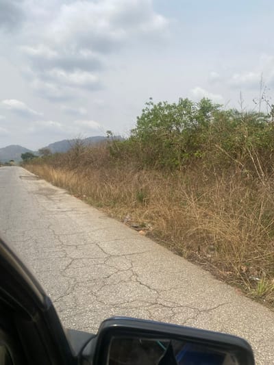 This image captures a degraded asphalt road, viewed from the interior of a dark-colored vehicle with its side mirror visible in the lower right, in Sasaro, Nigeria. The two-lane road, showing extensive cracking and minor potholes, extends towards the distant left and is bordered on the right by a strip of dry, tall, light brown grass and sparse green shrubs. Scattered plastic debris and other refuse are visible within this roadside vegetation. In the background, under a heavily overcast sky with light and dark grey clouds, a series of hazy, vegetated hills or mountains rise. No individuals are present in the scene.