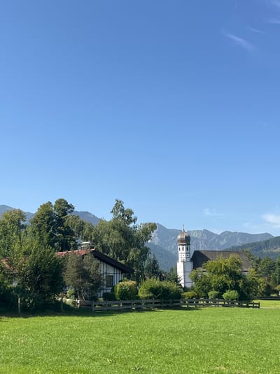 This image depicts a rural landscape in Fischbachau, Germany, on a clear, sunny day. The foreground features a vibrant green, mown grass field, with a low wooden fence partially visible extending from left to right. In the midground, a traditional Bavarian-style house with a white facade, dark timber accents, and a reddish-brown roof is situated on the left, partially obscured by various trees and dense green bushes. To the right, a prominent white church building with a distinctive onion-domed steeple (Zwiebelturm) and a dark-pitched roof stands partially enveloped by trees. The background is dominated by large, forested mountains under a clear, bright blue sky with minimal high-altitude cirrus clouds visible in the upper right. No individuals or specific actions are discernible.